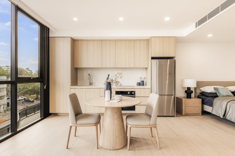 Arte Living Caringbah furnished studio with kitchen, dining table and queen bed beside floor-to-ceiling windows